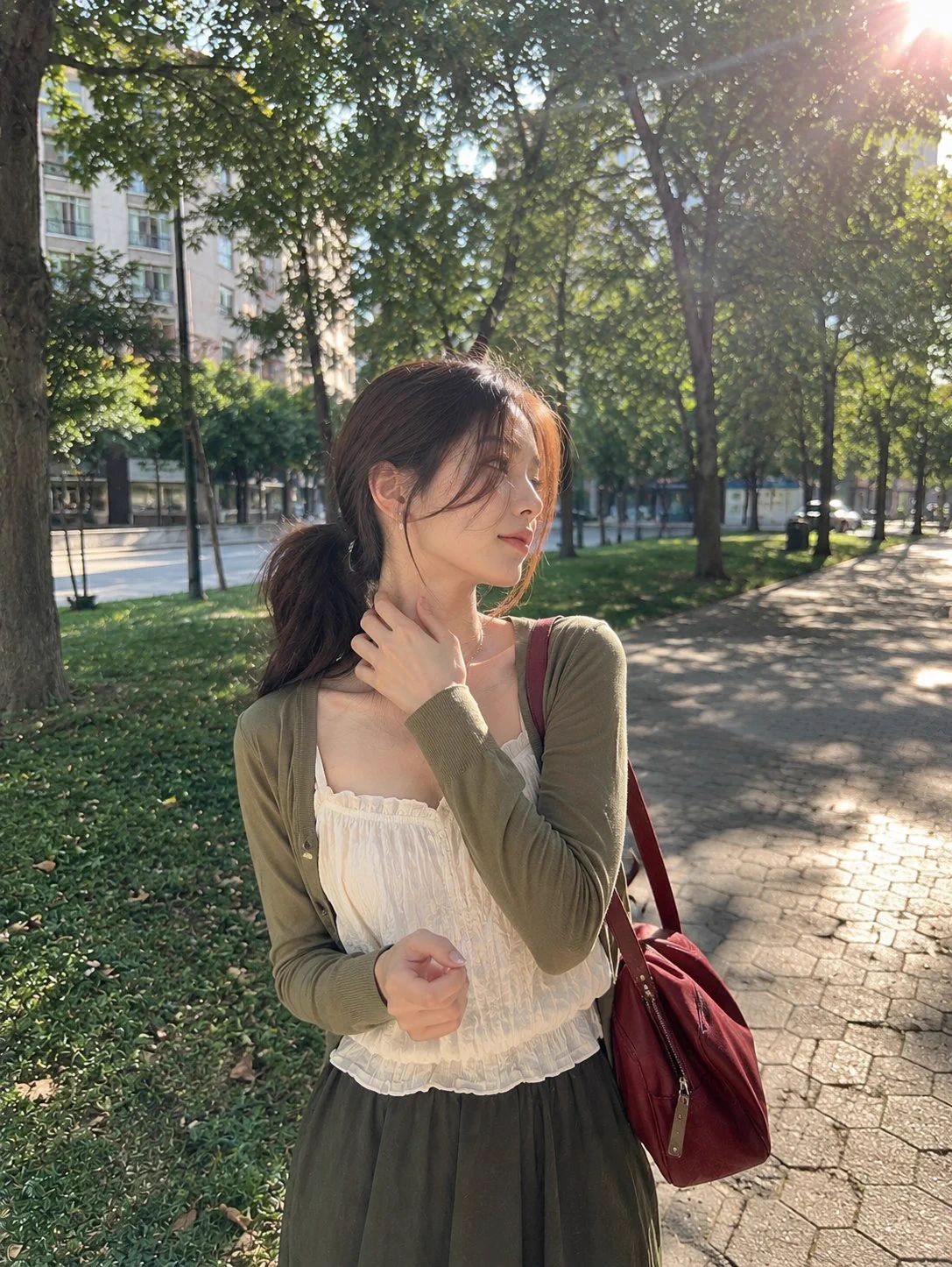 Femme en cardigan vert et sac bordeaux dans un parc ensoleillé.