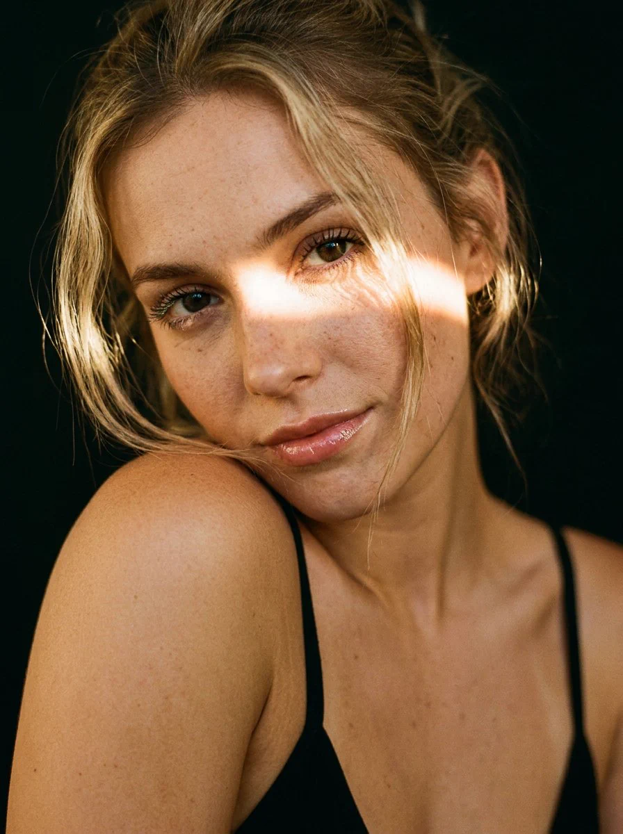 Gros plan d’une femme en débardeur noir, la tête inclinée, avec une bande de soleil chaud sur le visage et un fond sombre.